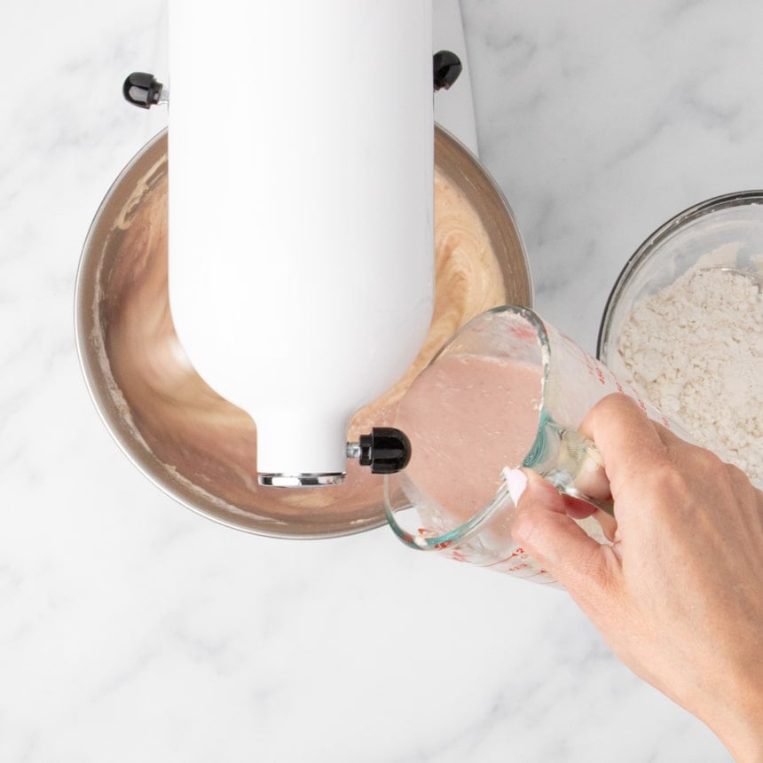 Hand pouring pink liquid into a mixing bowl of stand mixer while blending batter for strawberry cake