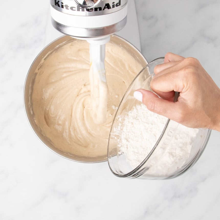 Hand adding flour mixture to smooth cake batter in a stand mixer bowl.