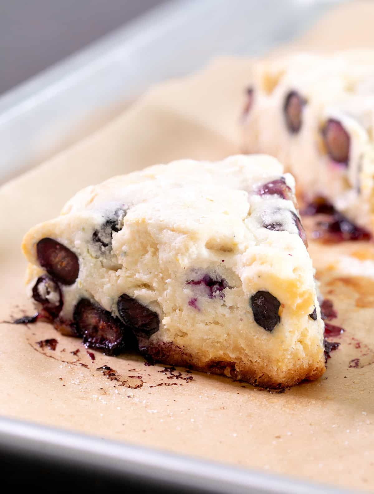 Closeup image of a light yellow baked triangle shaped pastry with blueberries on brown paper on a metal baking tray