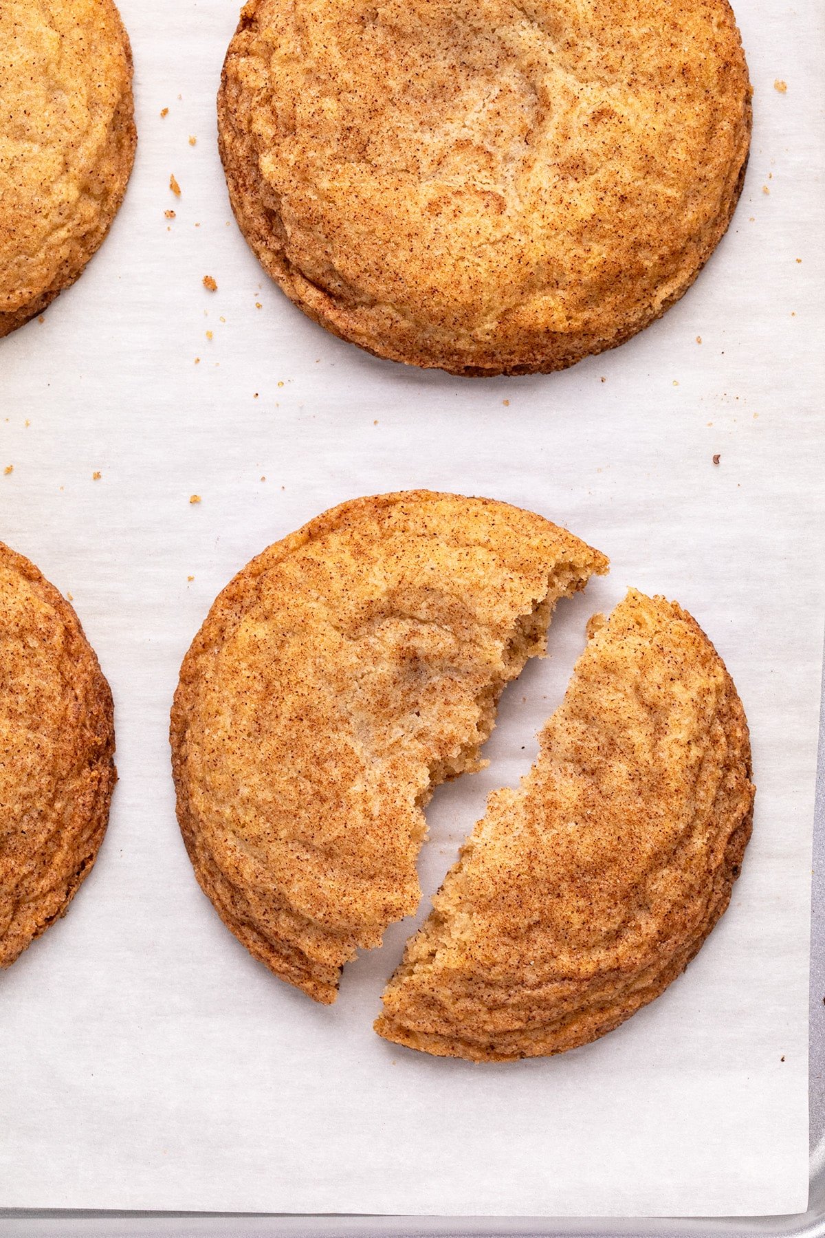 4 round crisp-edged brown cookies coated in cinnamon-sugar mixture on white paper lining baking sheet, with center cookie broken in half down the middle