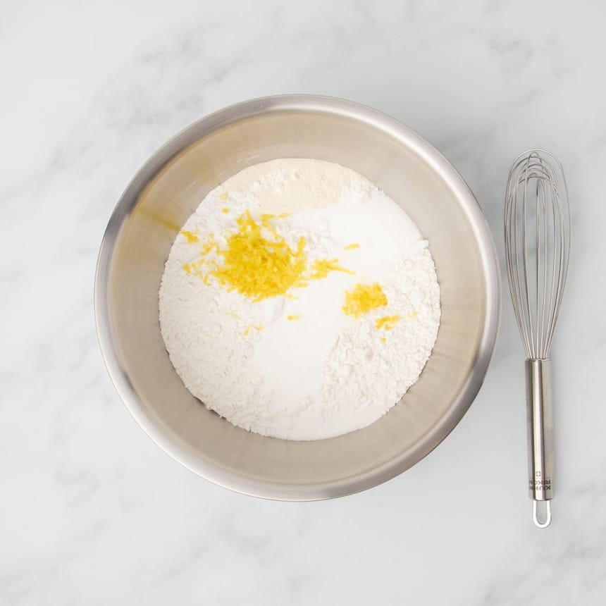 Large bowl with dry ingredients including flour, sugar, and yellow shredded lemon zest, with a whisk resting next to the bowl.