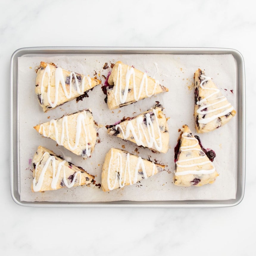 Freshly baked blueberry scones with a generous white glaze drizzled over them, on a parchment-lined baking sheet.
