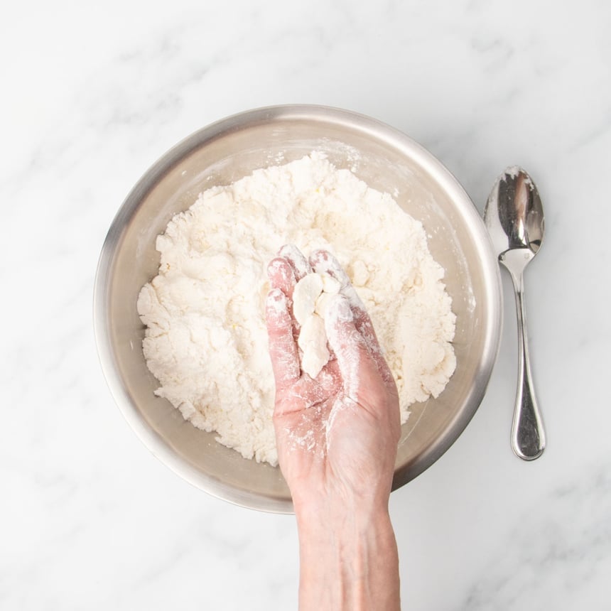 A hand holding clumps of butter and flour mixture, showing how to "cut" the butter into the dough by pressing the clumps flat.