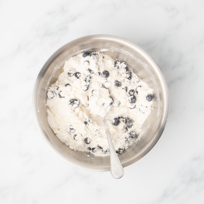 A bowl of dry ingredients and blueberries mixed with a spoon.