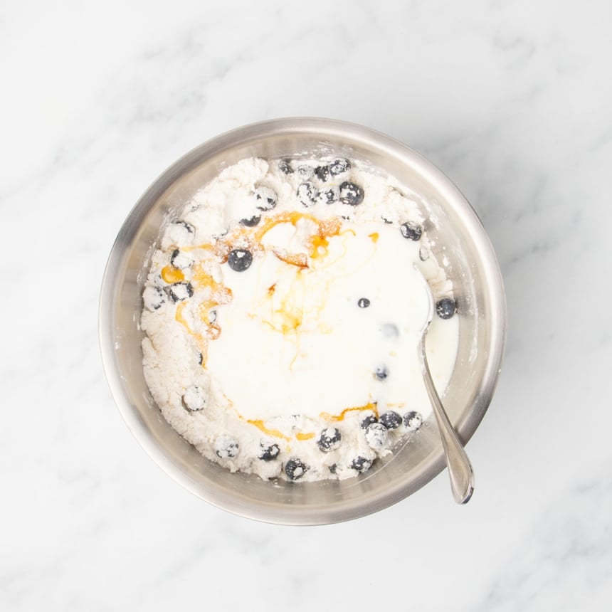 A bowl showing milk, honey, and vanilla poured over the flour, butter, and blueberry mixture.
