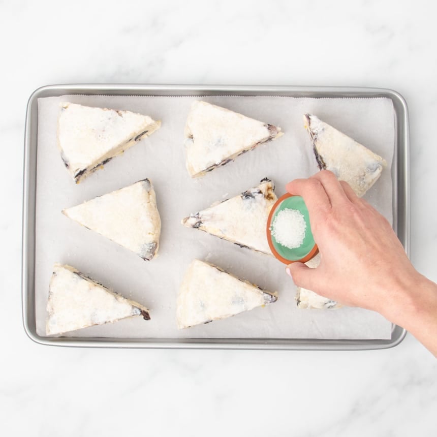 A hand sprinkling sparkling sugar over the unbaked dough wedges covered with cream on top, on a baking sheet lined with parchment paper.