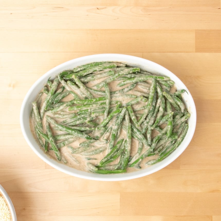 same green beans, sauce and grated cheese in white porcelain oval baking dish on blond wood surface
