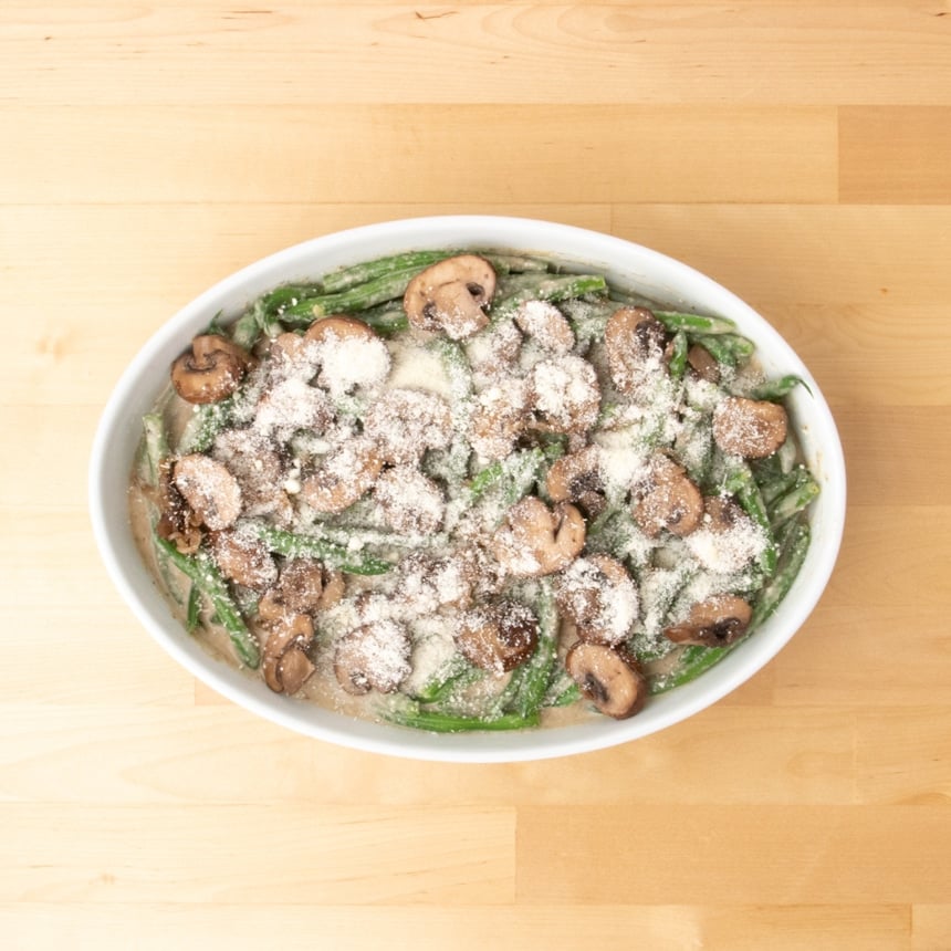 white oval casserole dish with green bean mixture now covered with a layer of brown sliced and cooked mushrooms and a sprinkling of white finely grated cheese