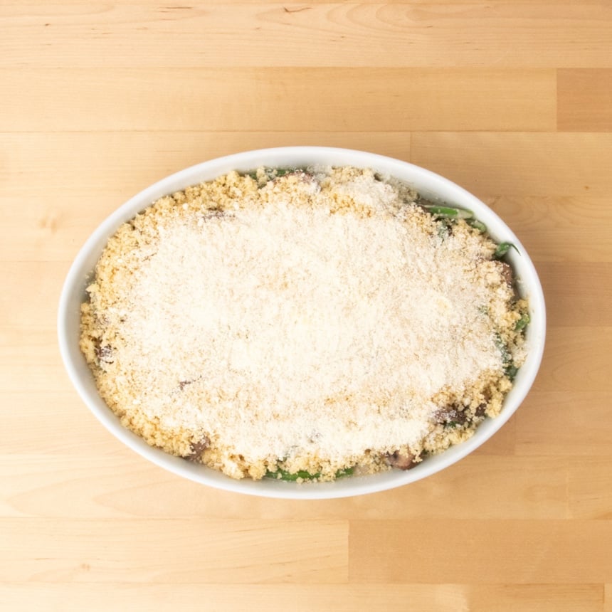 same casserole dish with the prepared breadcrumb mixture and a top layer of white grated cheese, ready to go in the oven