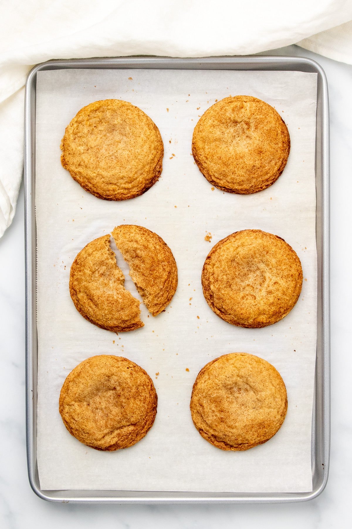 6 large, golden brown snickerdoodles, with one cookie broken in half, on white paper lining gray baking sheet with a white linen cloth at one short end