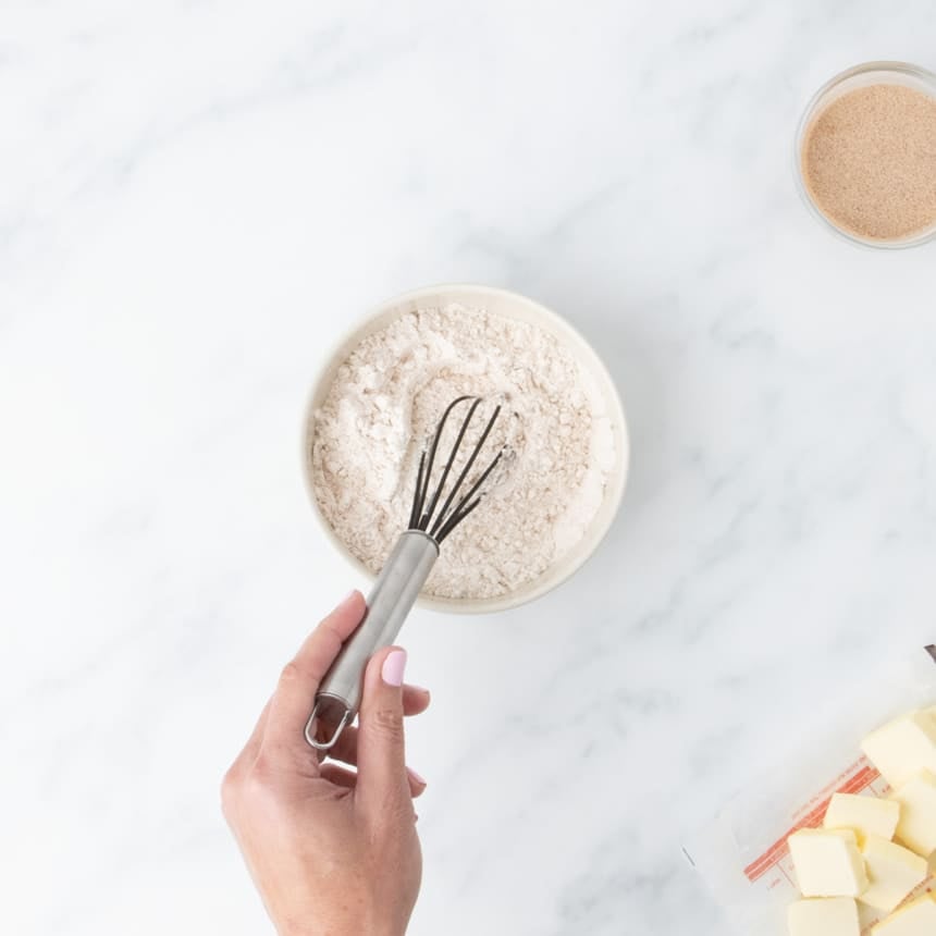hand holding small balloon whisk in small bowl of dry white flour mixture on marble surface