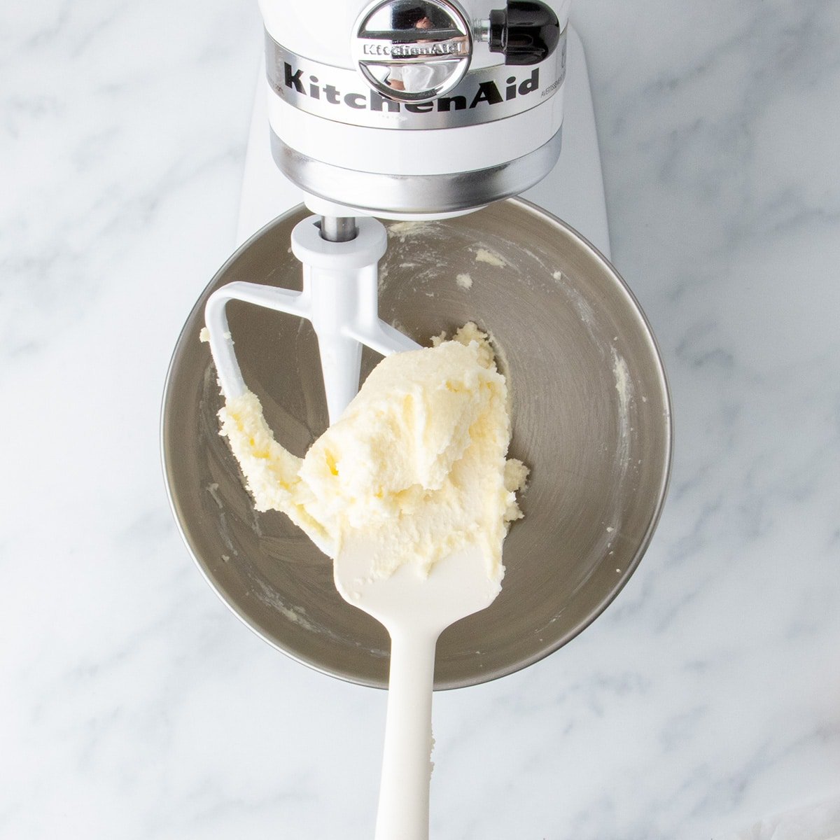 A white silicone spatula with pale yellow beaten butter and sugar mixture held over metal stand mixer bowl with white paddle attachment.