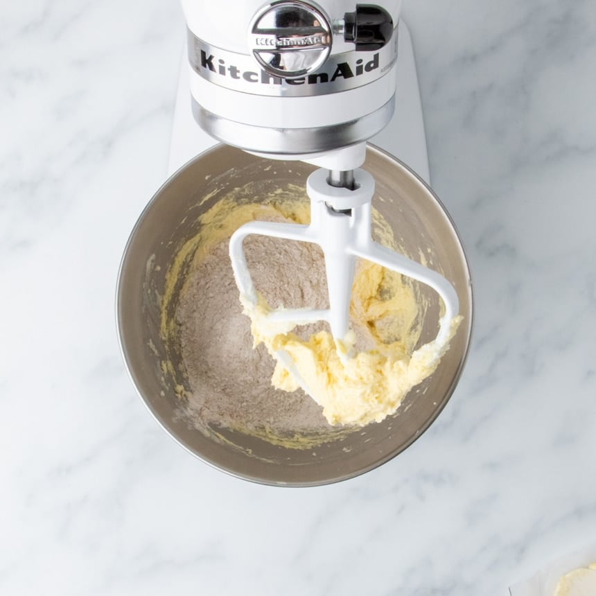 metal mixing bowl in stand mixer with egg, butter and sugar mixture on lifted paddle, and very light brown whisked dry ingredients on top of mixture in bowl