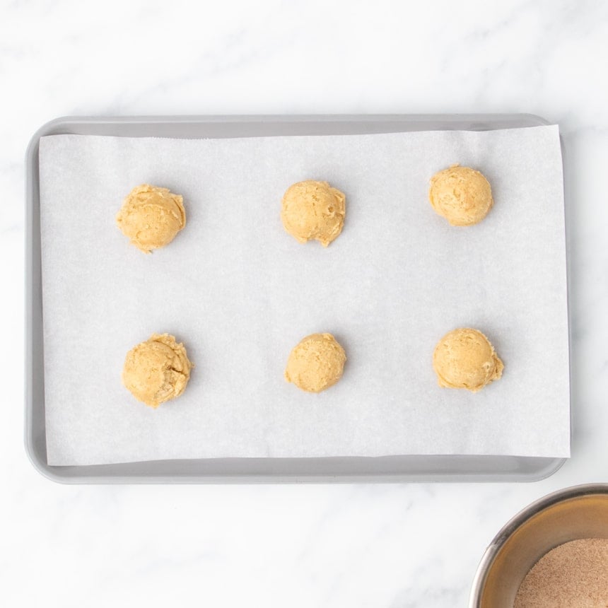 6 scooped portions of raw snickerdoodle cookie dough evenly spaced on white paper lining gray baking sheet on marble counter