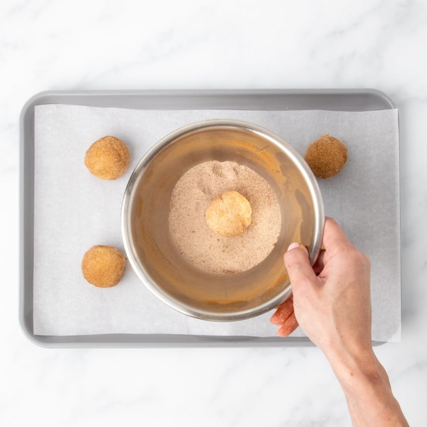 hand holding small metal bowl with cinnamon sugar mixture, covering ball of cookie dough in mixture