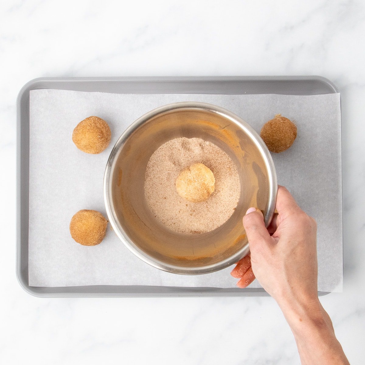 A hand holding small metal bowl with cinnamon sugar mixture, covering ball of cookie dough in mixture.