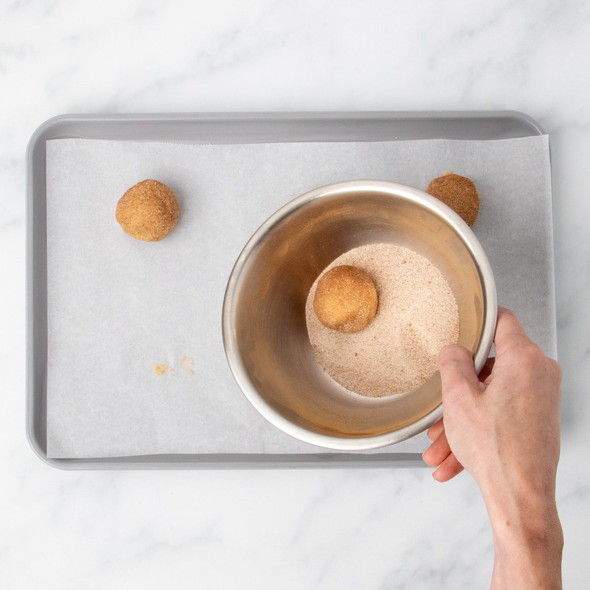 A hand holding a bowl with cinnamon sugar mixture, covering one raw ball of dough in mixture.