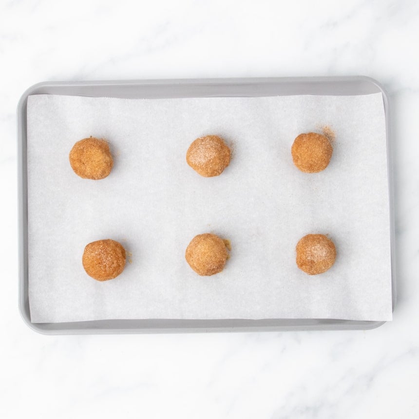 6 balls of raw snickerdoodle cookie dough, well-coated in cinnamon sugar mixture, on baking sheet ready to be baked