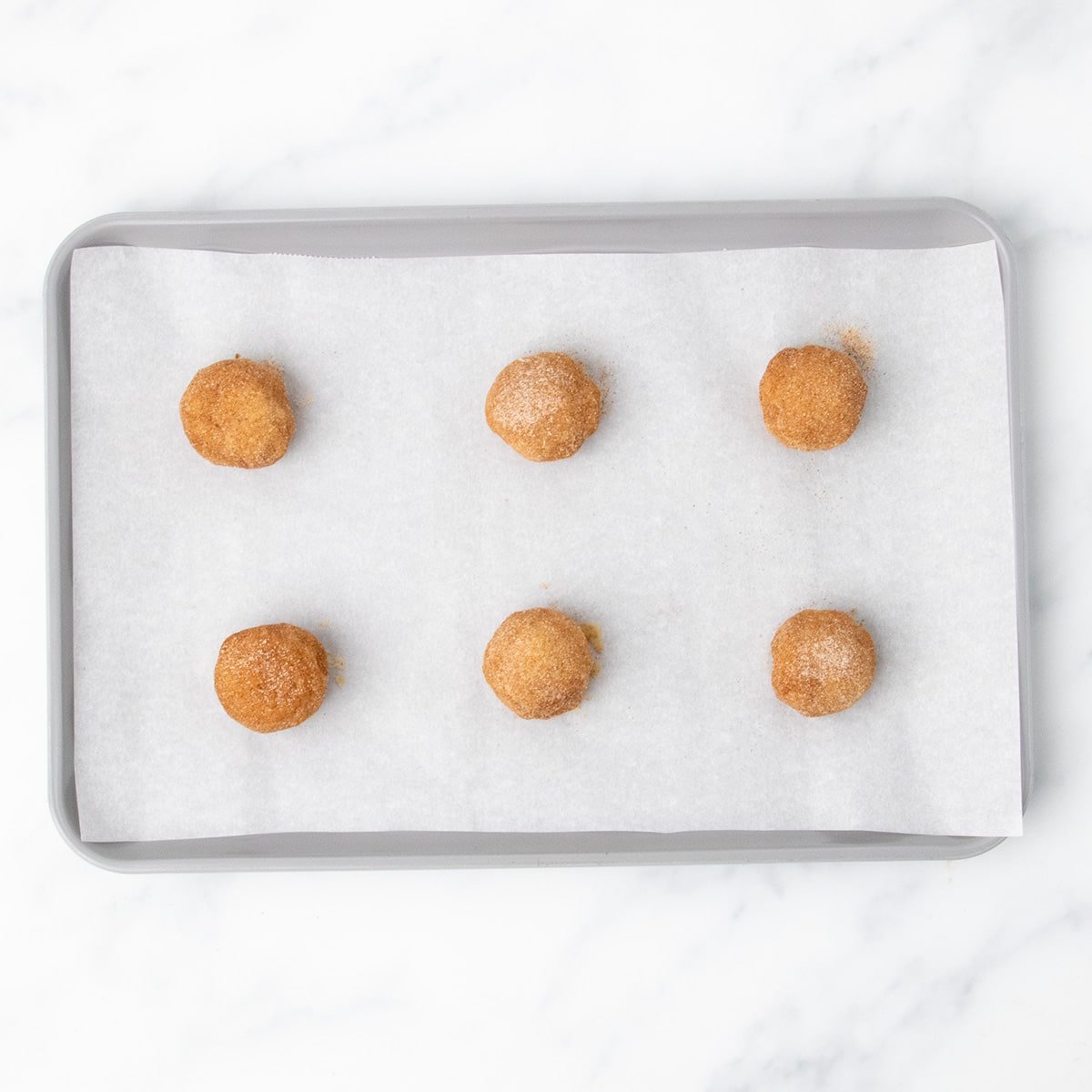6 balls of raw cookie dough, well-coated in cinnamon sugar mixture, on baking sheet ready to be baked.