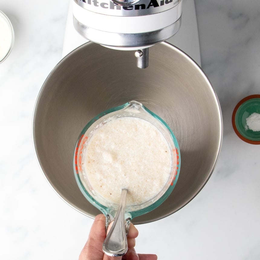 glass measuring with cloudy milk mixture with metal handle of spoon held over round metal stand mixer mixing bowl