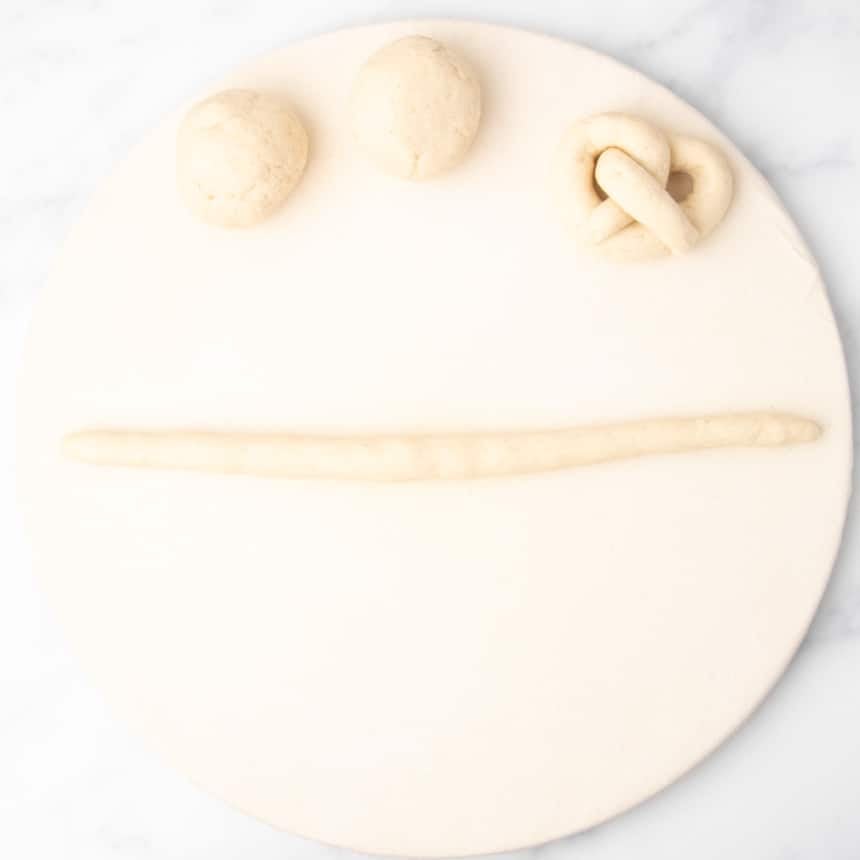 4 pieces of raw bread dough, 2 shaped into loose rounds, one in a pretzel shape, and one rolled out into a 12-inch long rope