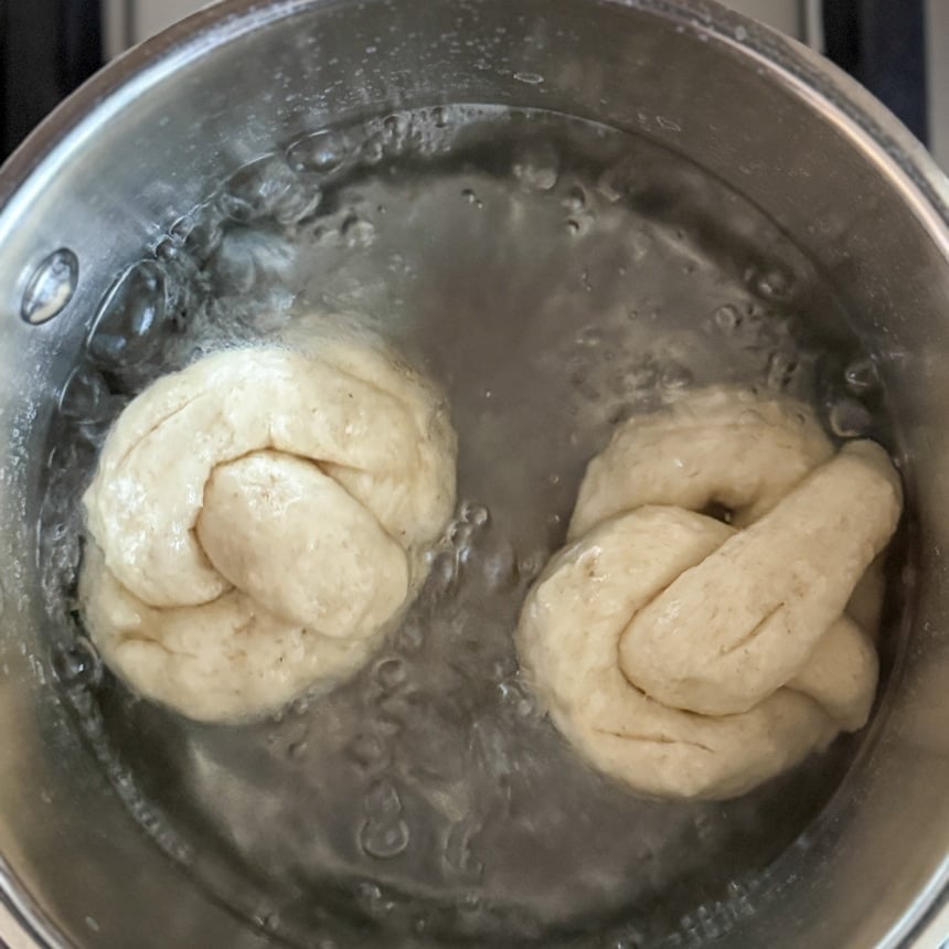 2 raw soft pretzel shaped pieces of dough boiling in baking soda bath