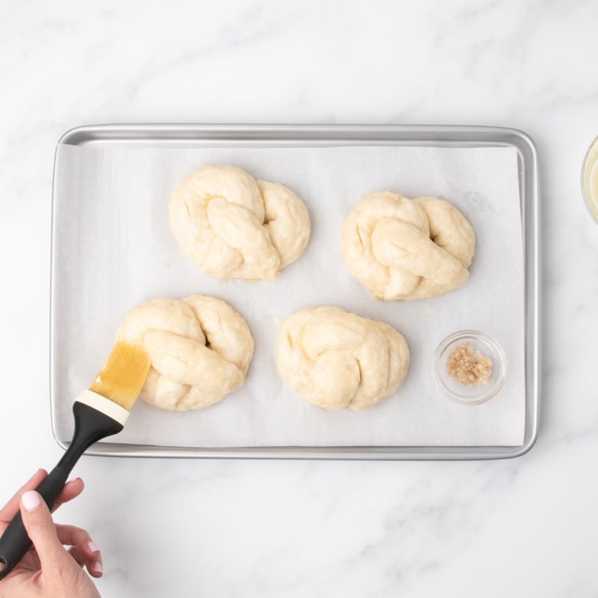 4 shaped, risen and boiled raw soft pretzels being brushed with melted butter with pastry brush, with coarse sea salt in a small bowl on tray