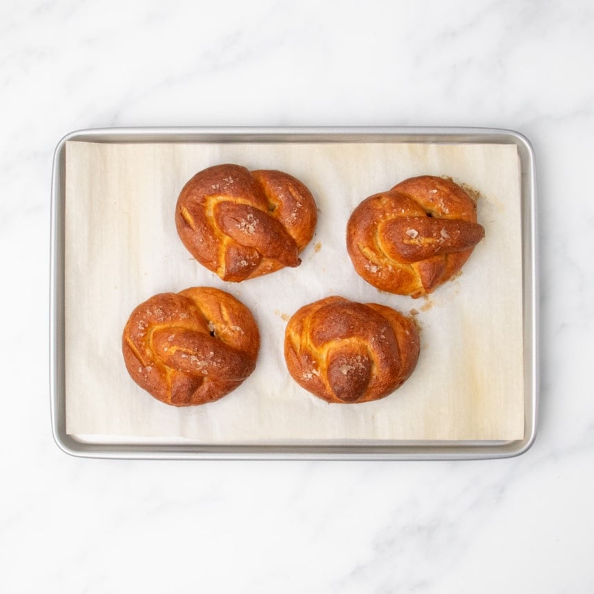 4 fully baked, golden brown gluten free soft pretzels with flaky sea salt on top, all on white paper lining on metal rectangular rimmed baking sheet