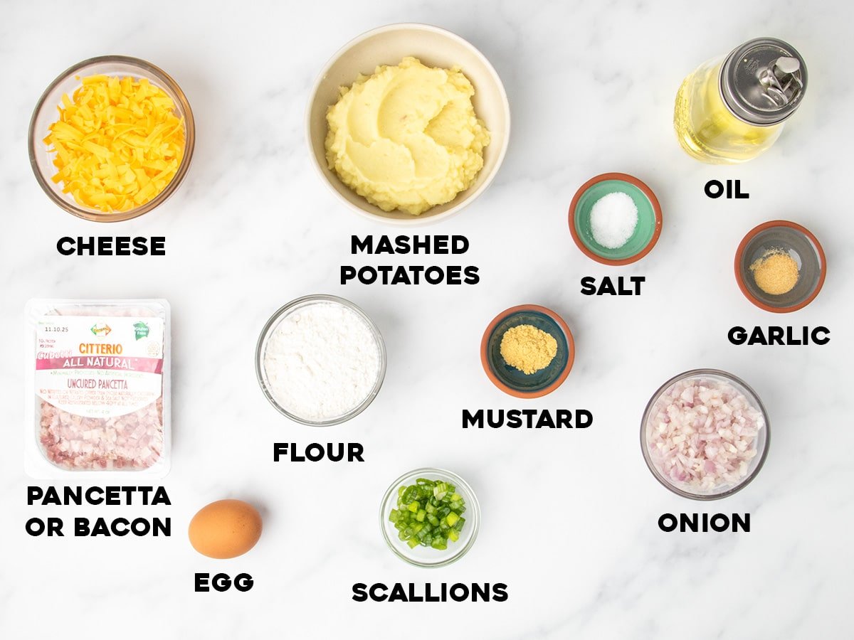 Bowls and small dishes of labeled ingredients arranged on a marble surface, including cheese, mashed potatoes, oil, diced pancetta, flour, salt, mustard powder, garlic powder, diced onion, egg, and scallions.