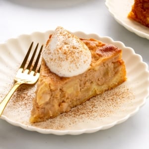 Closeup image of slice of cake with visible apple pieces on a white scalloped plate topped with whipped cream and sprinkled with cinnamon, gold fork beside it.
