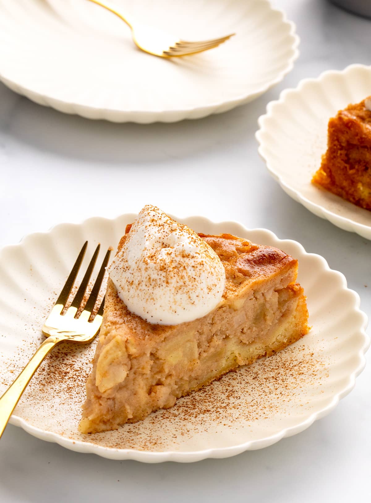 Slice of gluten free apple cake with whipped cream and cinnamon on a scalloped plate, surrounded by one small empty plates with a small gold forks and one plate with the back of another slice of cake.