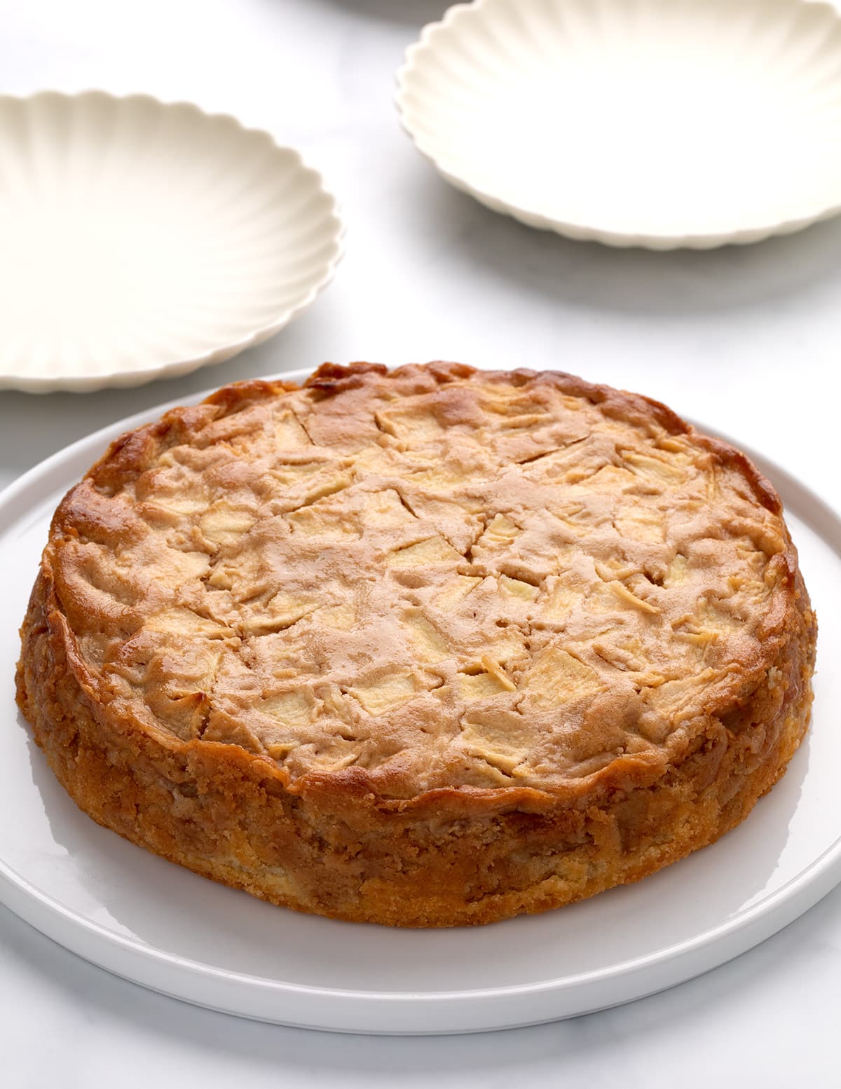 Whole round golden brown cake with visible cooked apple pieces on a white platter, small white cake plates in the background.
