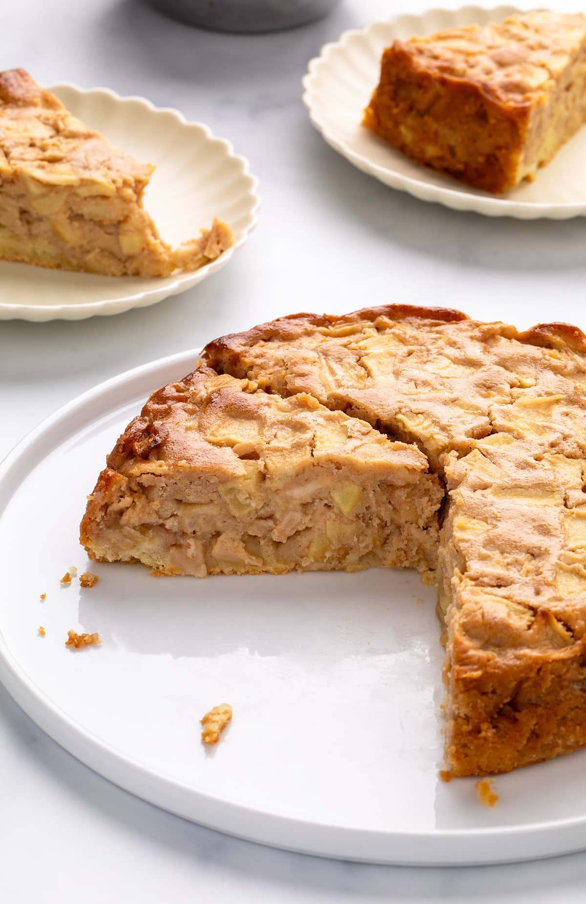 Round gluten free apple cake with a slice removed on a white platter, showing the moist interior with apple pieces; slices on plates in the background.