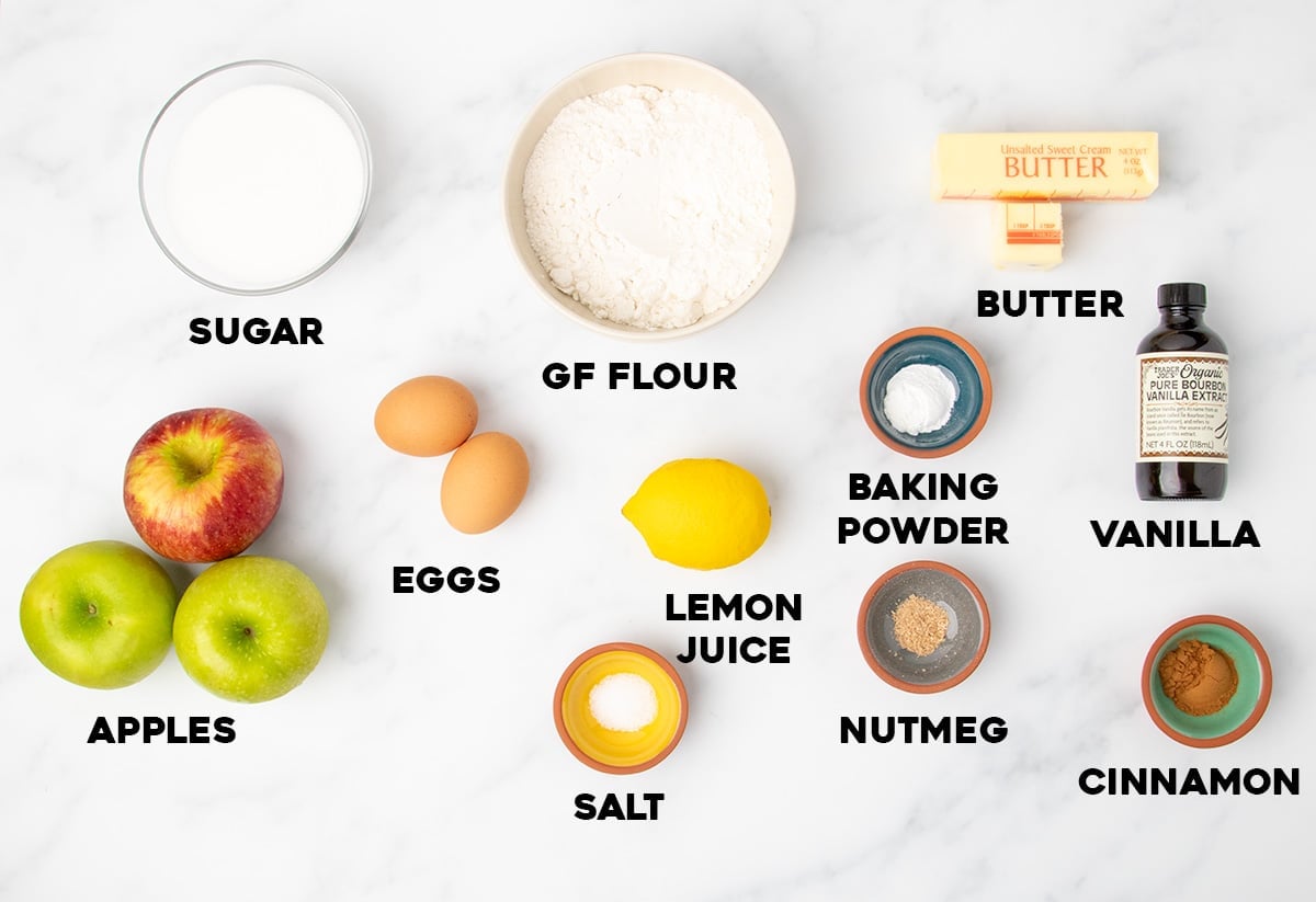 Overhead view of baking ingredients on a white surface labeled with text: sugar, gluten-free flour, butter, vanilla, baking powder, cinnamon, nutmeg, salt, lemon juice, eggs, and apples.