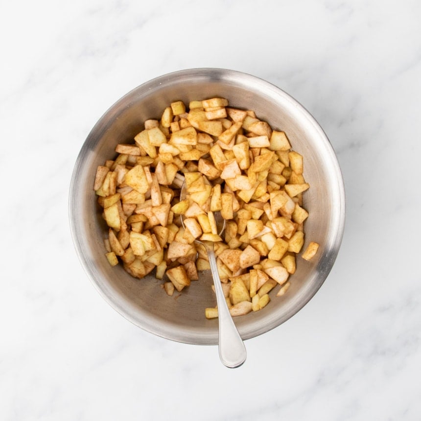 Small diced apple pieces coated in cinnamon mixture in a metal bowl with a spoon.