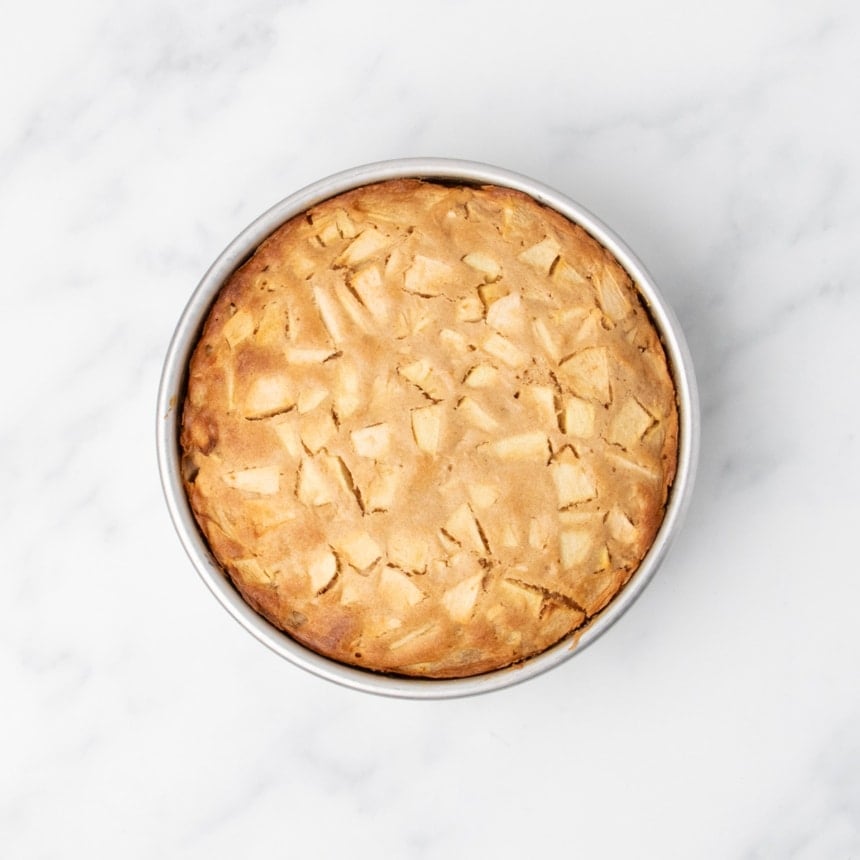 Baked apple cake in a round metal pan, golden brown on top with visible apple pieces.