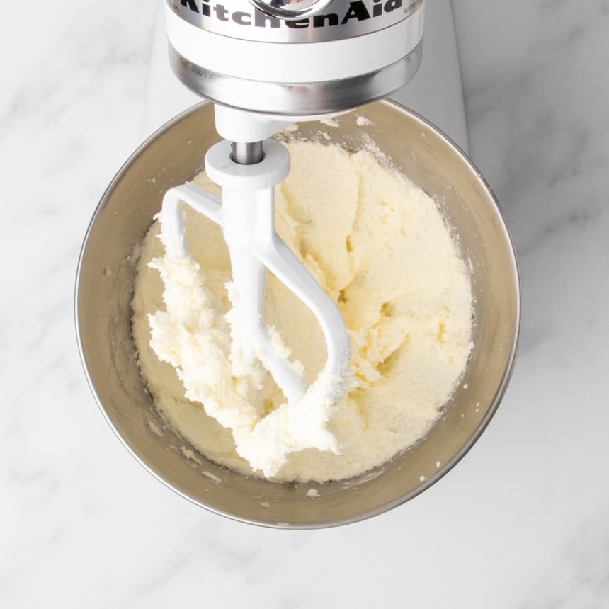 Butter and sugar mixture in stand mixer bowl after being creamed until fluffy, partly on white mixer paddle attachment