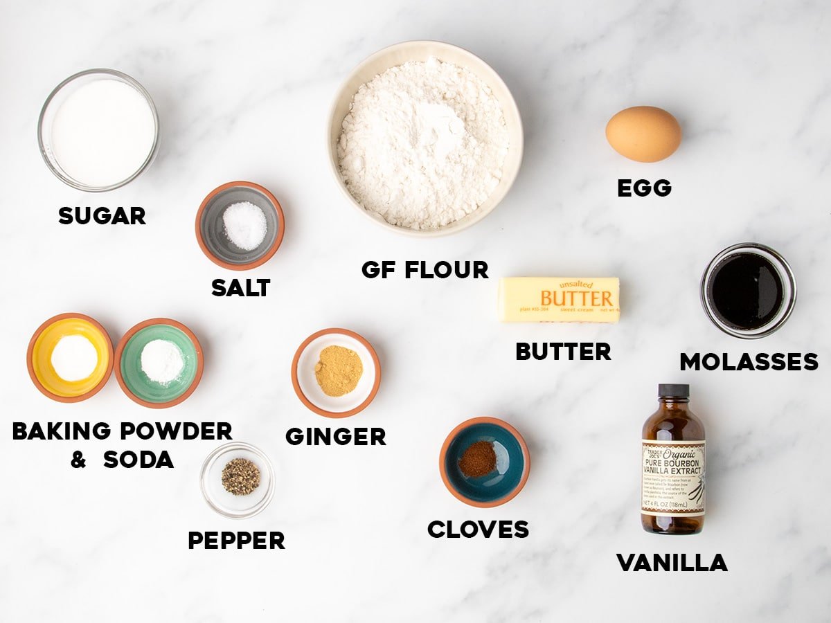 Overhead view of labeled baking ingredients in bowls and jars on a white surface, including gf flour, sugar, egg, butter, molasses, and spices.