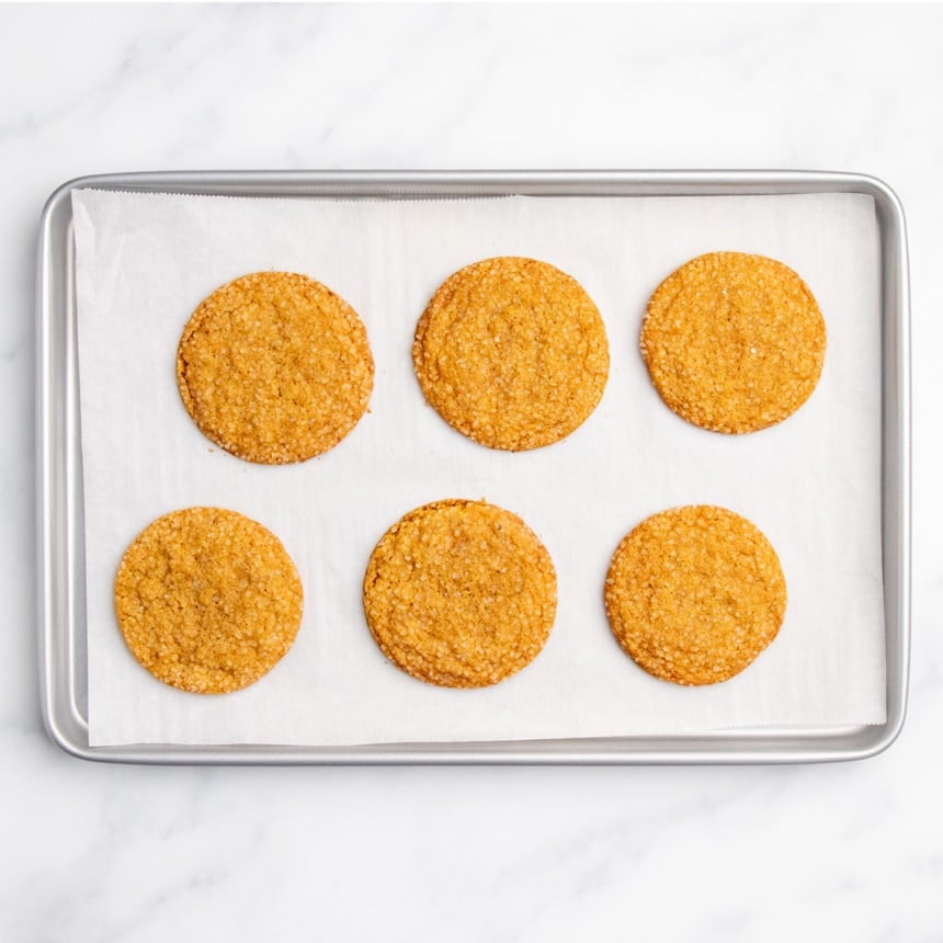 Six baked cookies with coarse sugar on top cooling on a parchment-lined baking sheet.