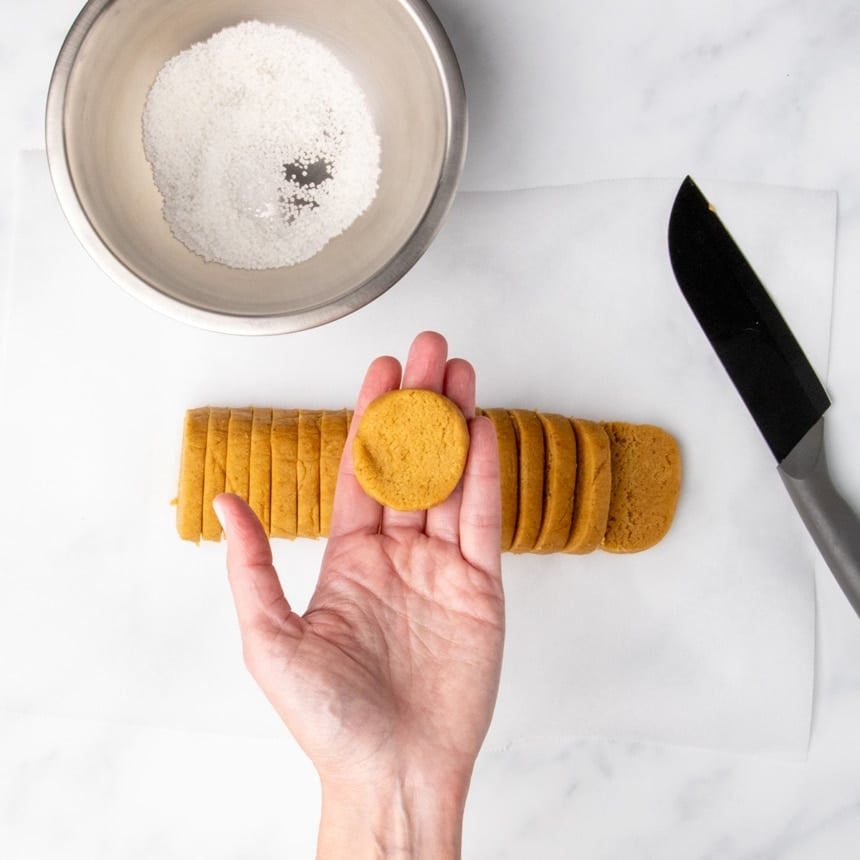 Hand holding a round slice of cookie dough with sliced dough and a bowl of coarse sugar in the background.