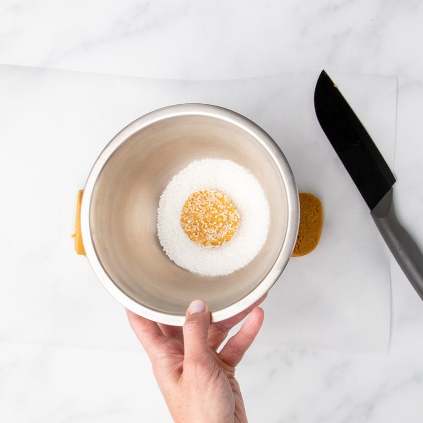 Hand holding a small stainless steel bowl with a slice of cookie dough coated in coarse sugar.