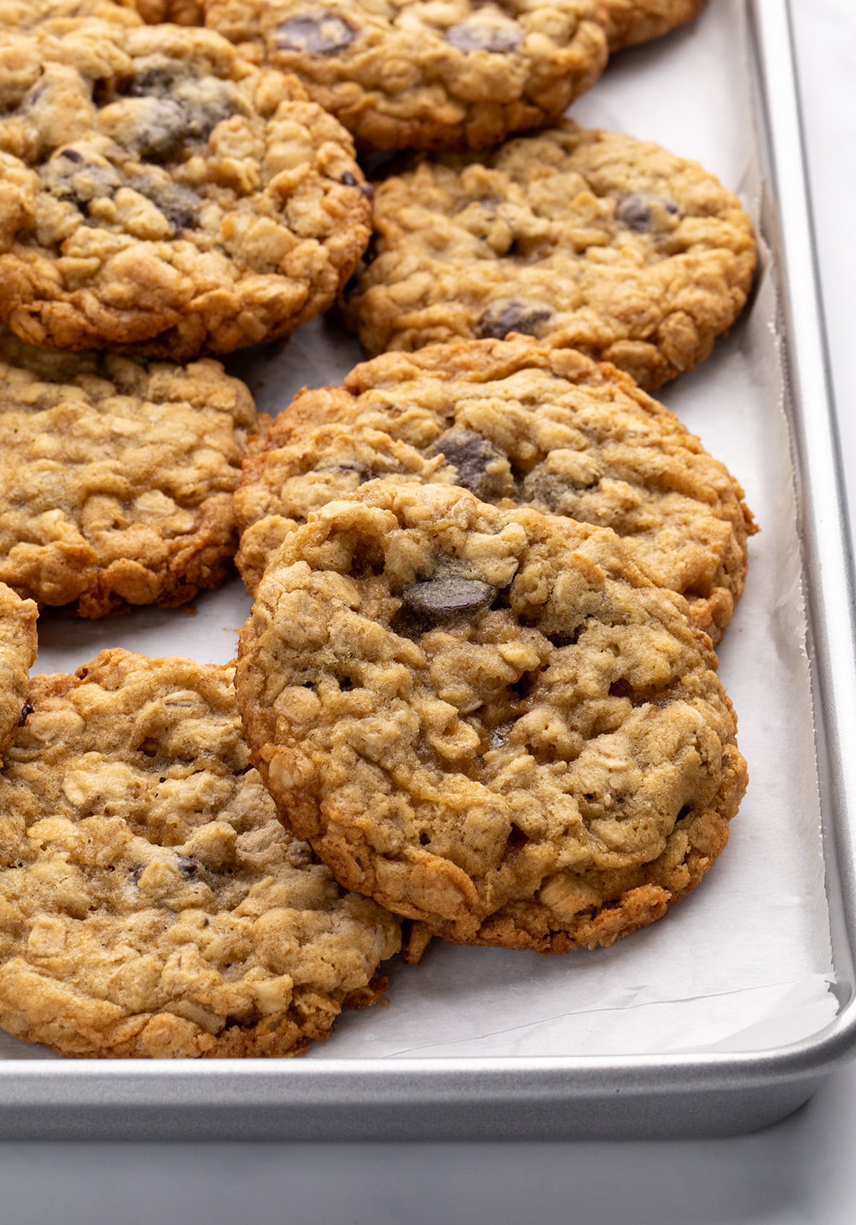 A closeup image of a corner of a metal baking sheet lined with white parchment paper, filled with evenly spaced oatmeal cookies with chocolate chips.