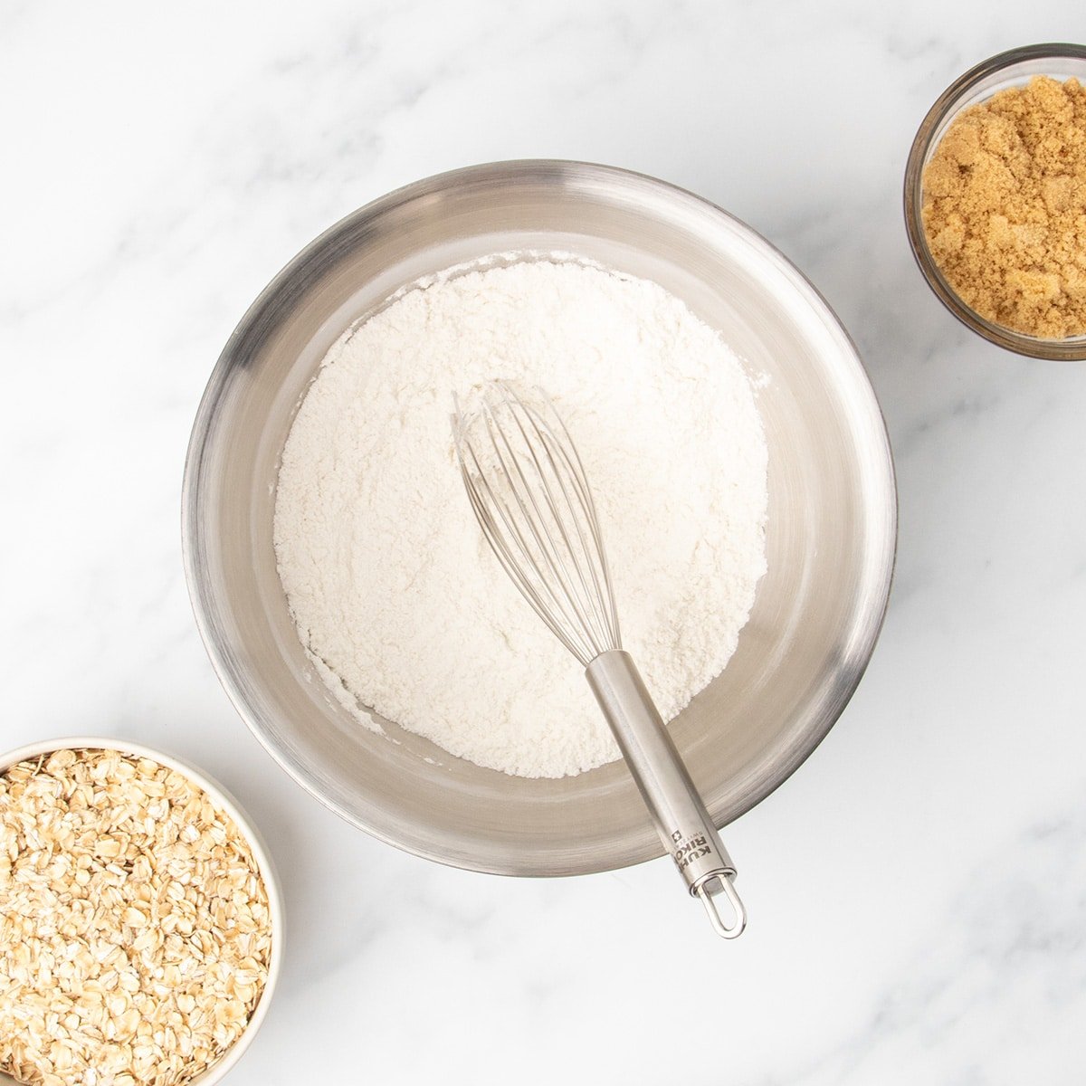 A metal mixing bowl filled with white gluten free flour and a whisk resting inside it, with a small bowl of rolled oats and a small bowl of brown sugar nearby on a white surface.