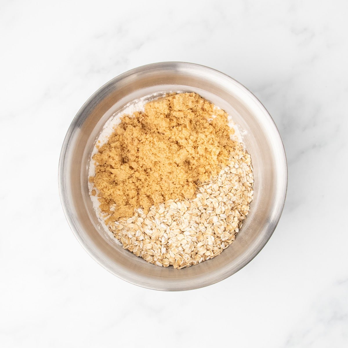 Same metal mixing bowl containing gluten free flour with brown sugar and rolled oats sitting on top, waiting to be mixed.