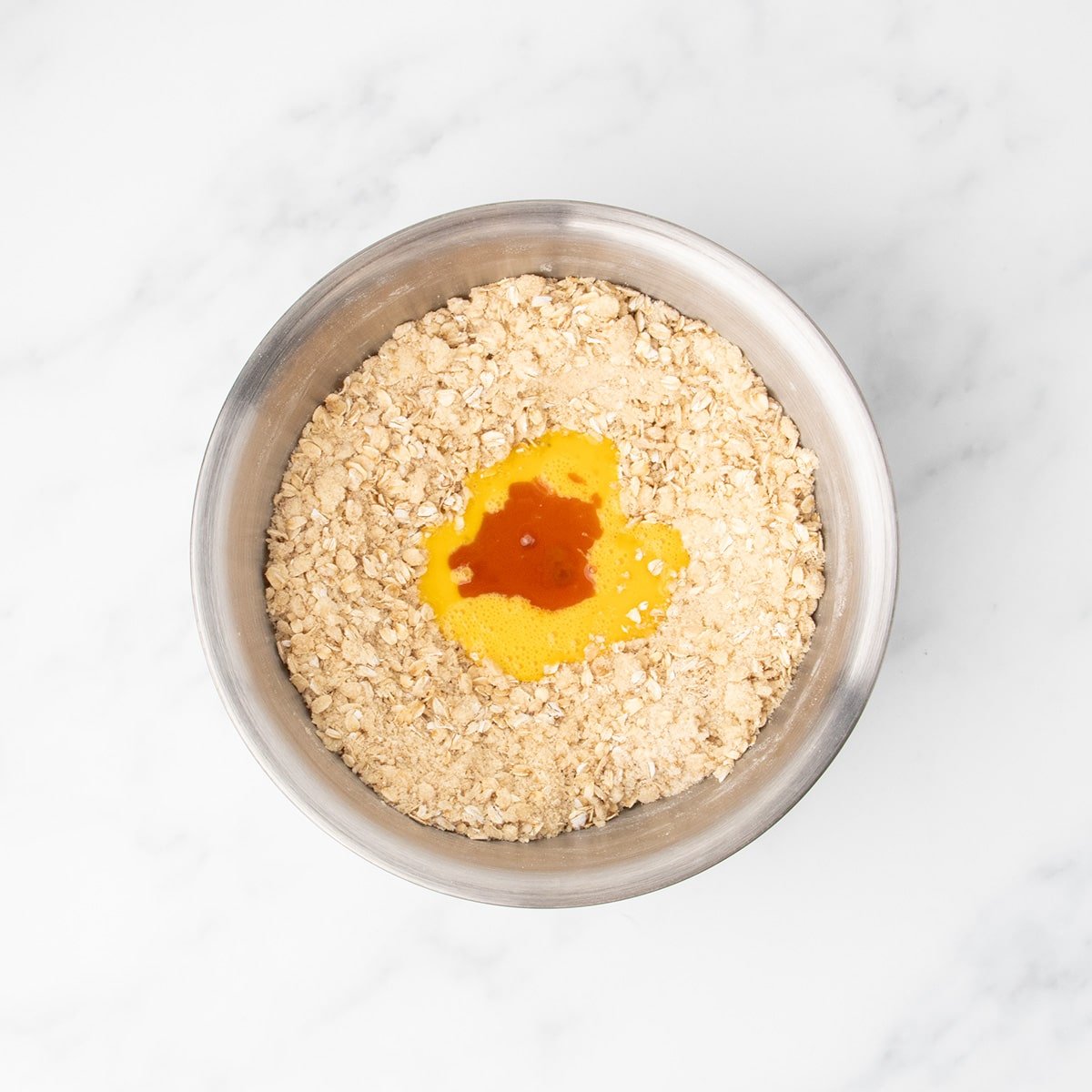 A metal mixing bowl with dry ingredients including oats, brown sugar, and fully incorporated softened butter, with a pool of beaten eggs and vanilla in the center waiting to be mixed.