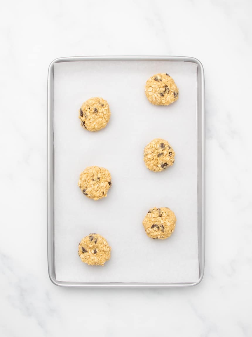 A metal baking sheet lined with white parchment paper, holding six evenly spaced unbaked disk-shaped mounds of oatmeal cookie dough.