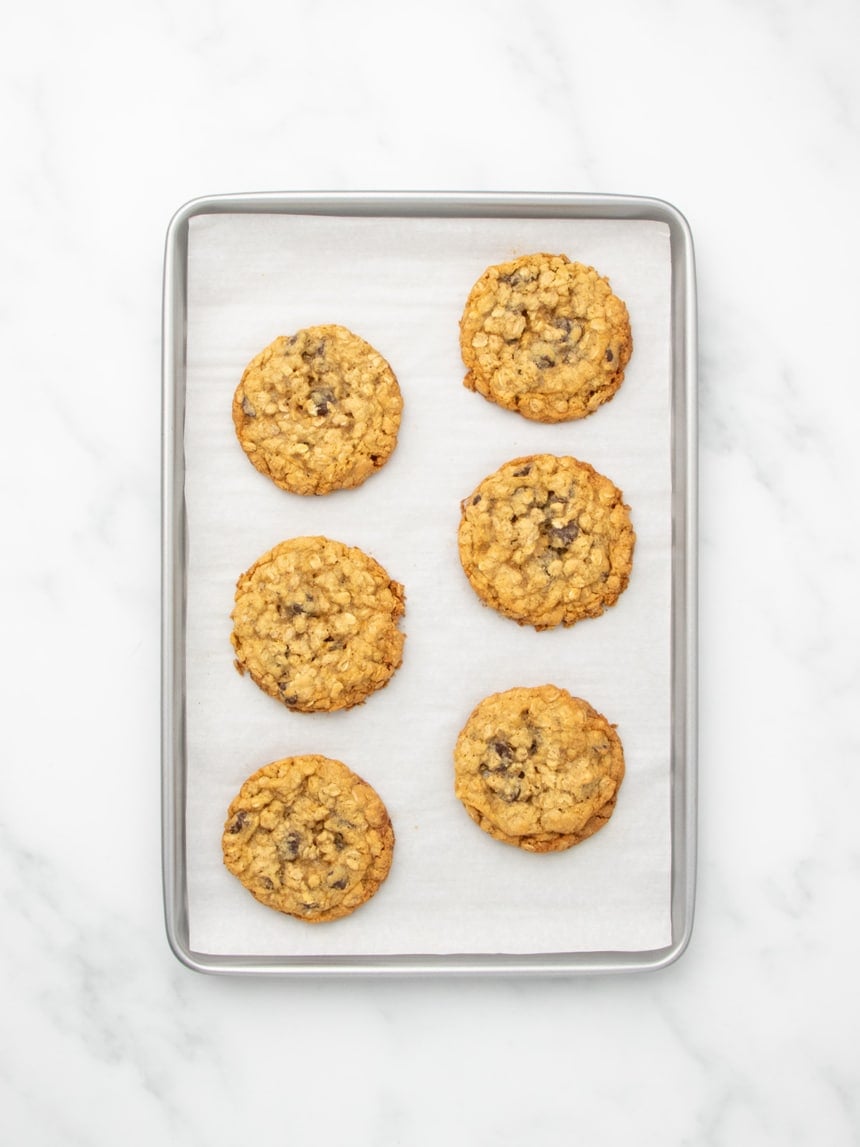 The same metal baking sheet lined with white parchment paper as in the previous photos, now holding six gluten free oatmeal cookies with chocolate chips baked to golden brown perfection.