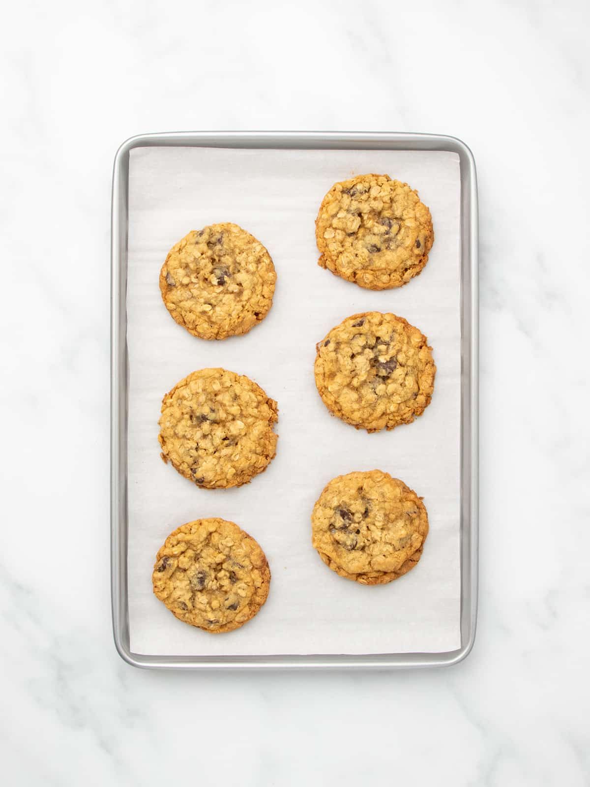 The same metal baking sheet lined with white parchment paper holding six gluten free oatmeal cookies with chocolate chips baked to golden brown perfection.
