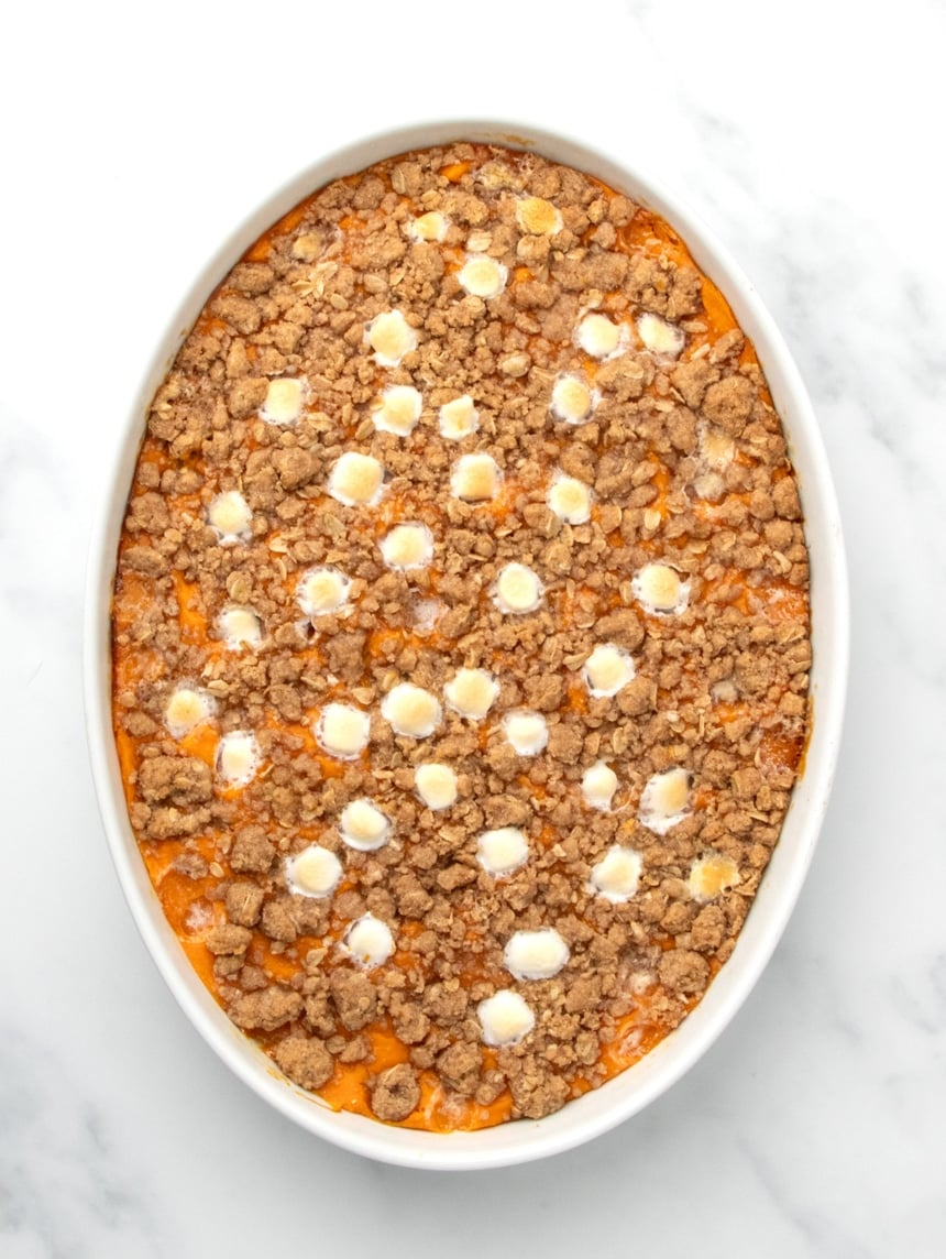 A white oval baking dish filled with orange sweet potato filling topped with a browned baked crumb mixture and melted marshmallows on a white marble surface.