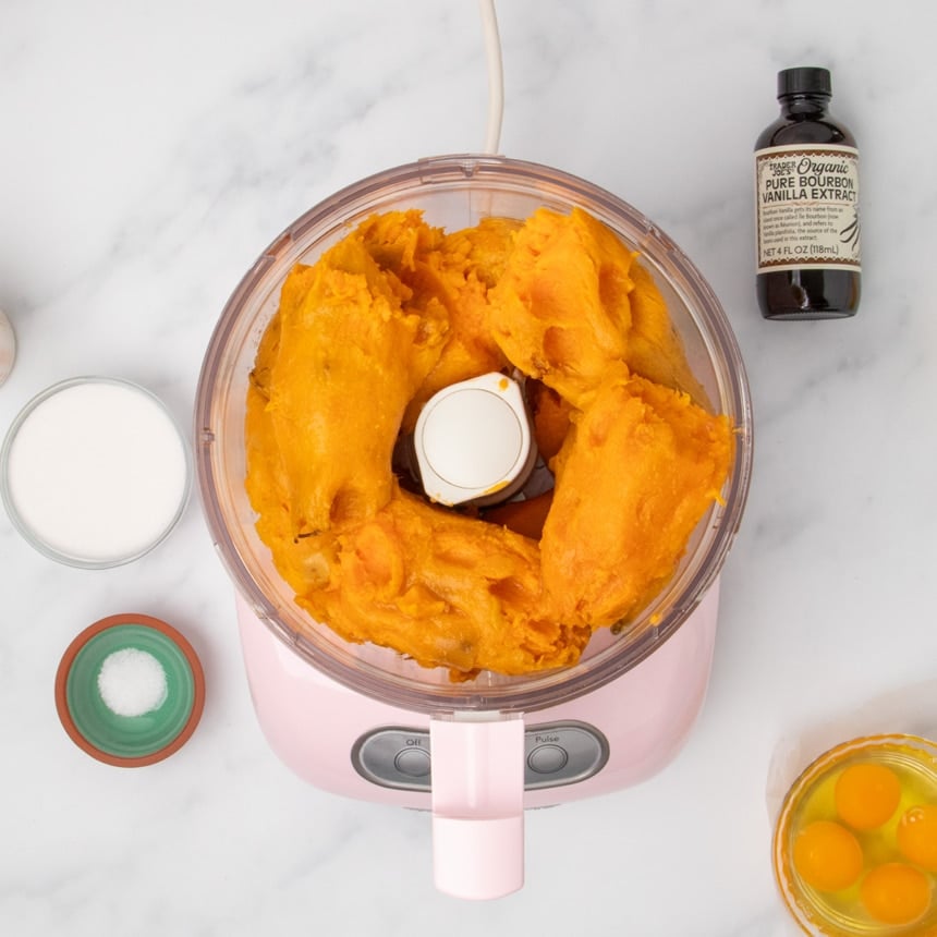 A food processor filled with cooked orange sweet potato pieces surrounded by small bowls of sugar, salt, 4 eggs out of the shell, and a bottle of vanilla extract on a white marble surface.